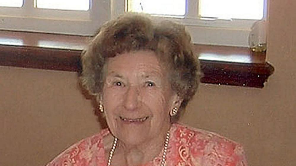 Woman with light-brown hair smiling. She is wearing a pink top, a pearl necklace and matching earrings.