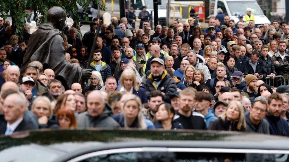 A large crowd of people mostly wearing black crowd into a street. A black car is in the foreground.