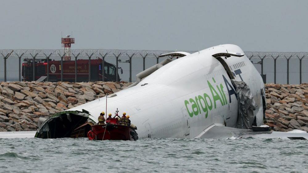 A damaged fuselage of a plane partially submerged in seawater. A small red boat carrying emergency workers is floating in front of the plane.