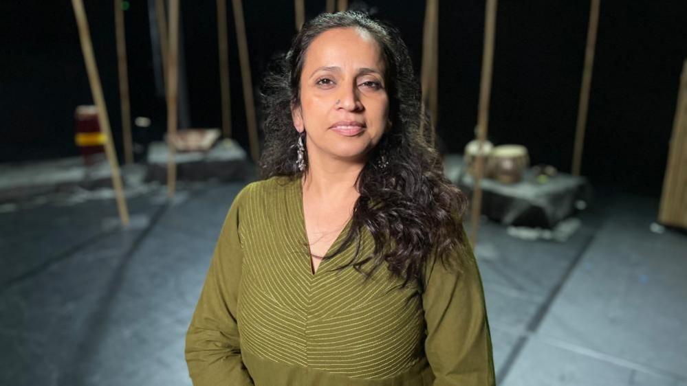 Urja Desai Thakore in a green outfit on a stage smiling into the camera. She has long, curly black hair and long earrings.