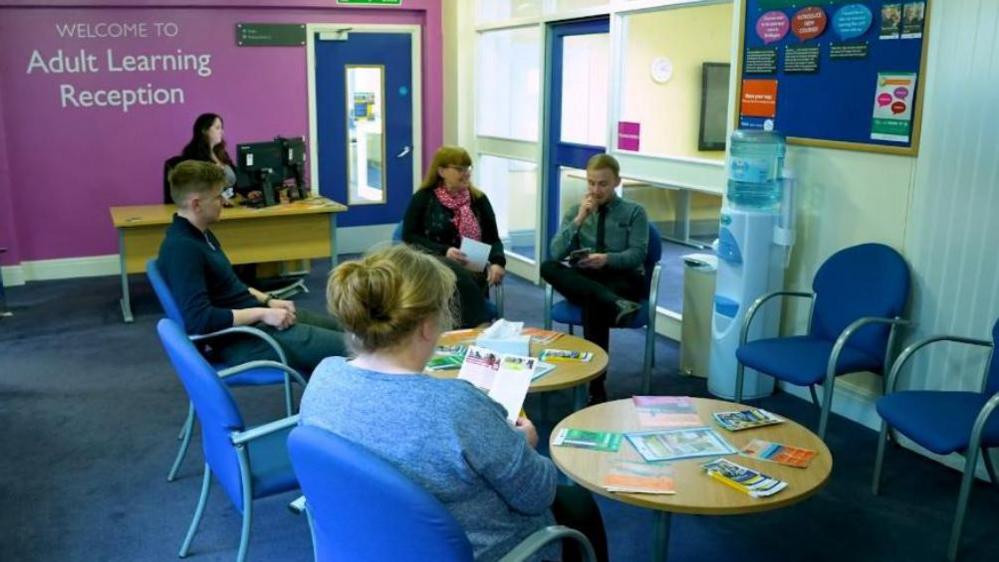 Two men and two women sit around two circular tables in an adult learning centre. One of the women is looking at a leaflet. 