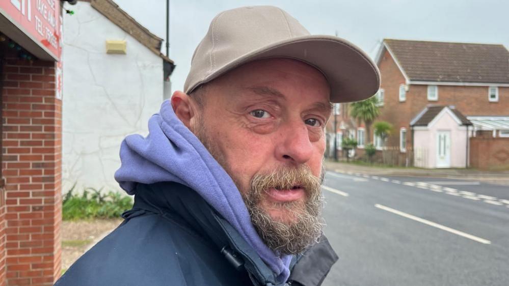 Ade Evans is bearded, and is wearing a blue jacket and light blue hoodie and a beige baseball cap. He is standing outside the Chinese take-away on Acle High Street.