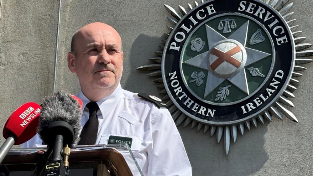 A man wearing a white PSNI shirt and black tie standing in front of microphones. There is a large PSNI logo on the wall behind him.