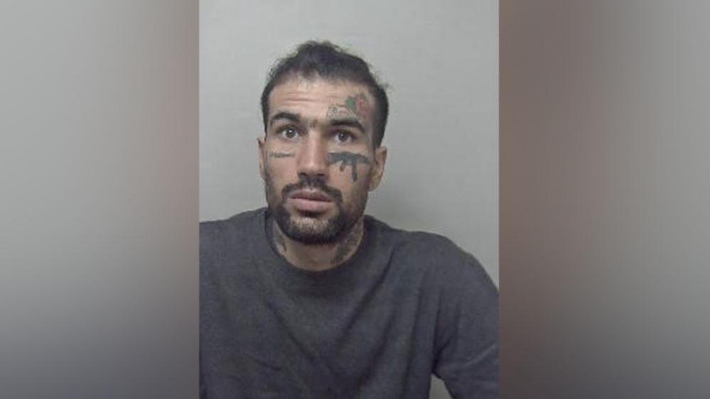 Custody photo of Fayaz Khan, who has short dark hair, a beard and moustache. He has tattoos on his face, including one of an AK-47 beneath his left eye. He is wearing a grey jumper