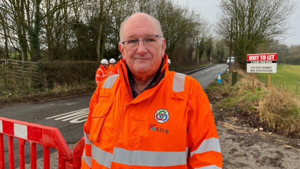 Phil Larratt with thin grey hair wearing orange high-visibility jacket next to barrier on a road