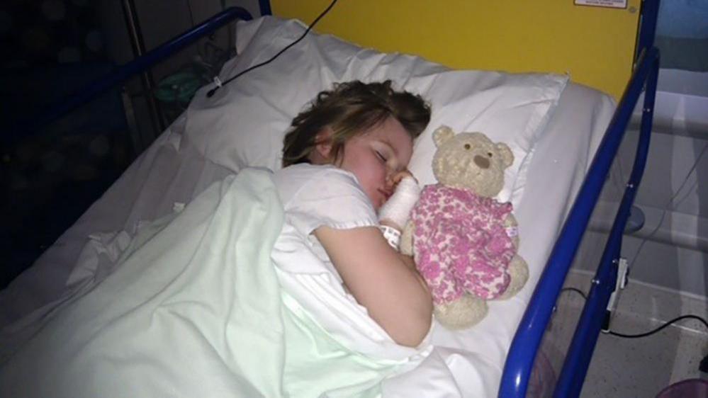 Jamilla Blake as a seven-year-old asleep on her side in a hospital bed with a teddy bear on the pillow beside her