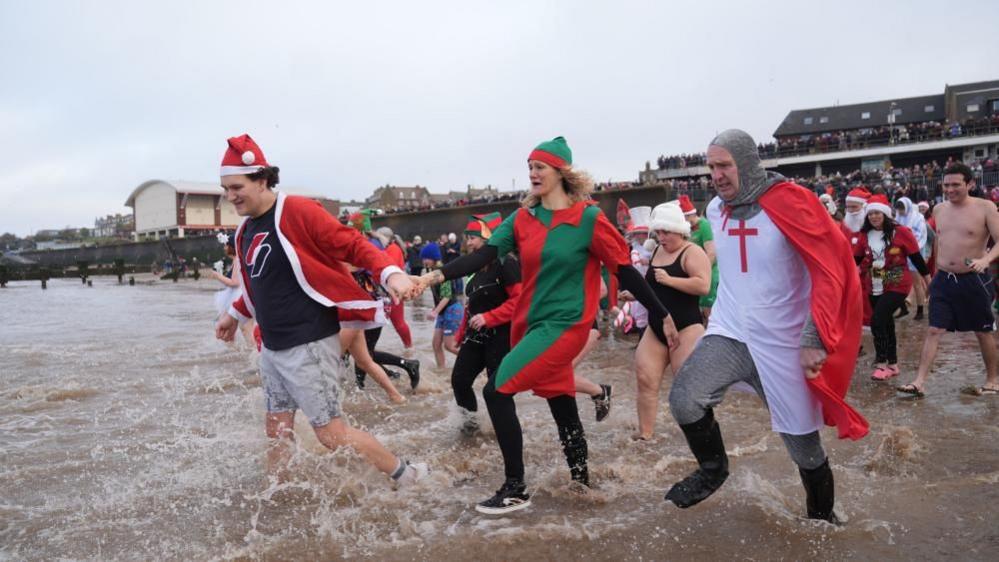 A man dressed as santa, a woman in a red and green dress and a man dressed as St george complete with pretend chainmail, go into teh sea as hundreds look on.