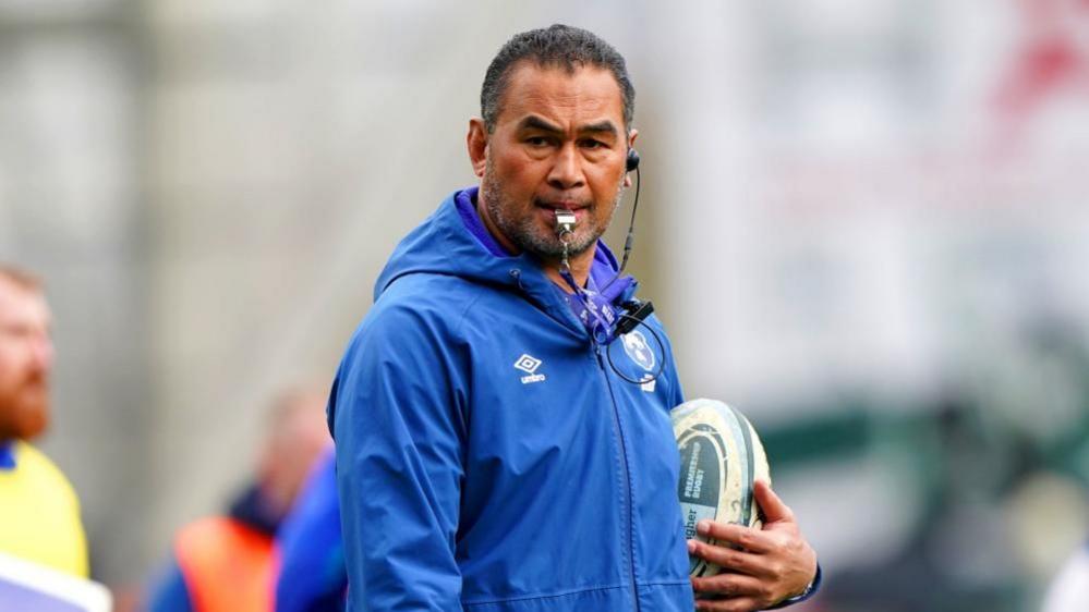Pat Lam, wearing a blue coat and holding a rugby ball. There is a whistle in his mouth.