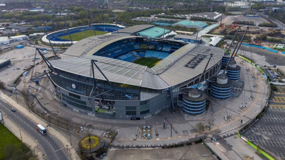A large football stadium see from above. It is surrounded by a carpark on one side, another football pitch on another and a road on another. It has the word Etihad printed on the top of the main stadium. 
