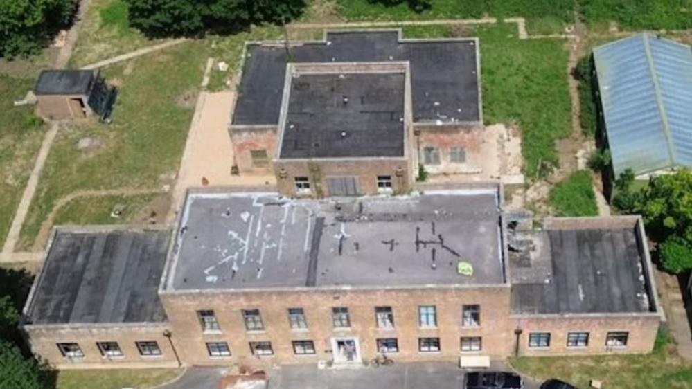 The former RAF West Raynham station headquarters building, is a 1940s brick built block which is symmetrical with a central entrance. The main block is two storeys high, with seven windows across the upper floor. That block is flanked by two single storey blocks which each have three windows on the front elevation. The building has a flat felted roof, and shows another block on a third storey behind, and a further ground floor block - and as outbuilding on either side of the image.
