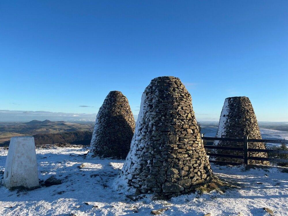 The Three Brethren cairns, which consist on three tall mounds of stones, outside Selkirk, with Eildon Hill visible in the background. It's a snowy day with a blue sky.