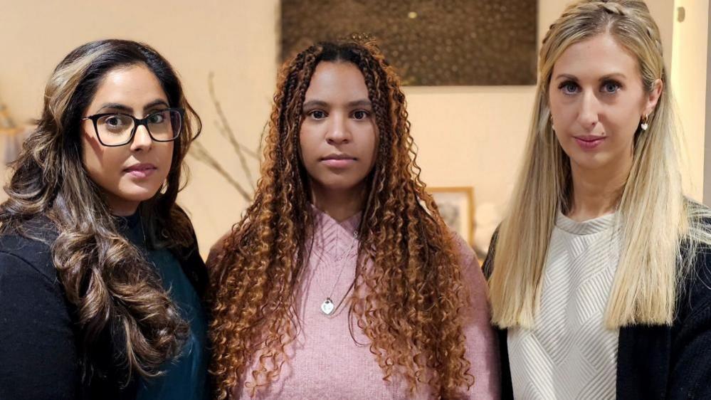 Three women stand side-by-side with serious expressions on their faces. The woman on the left wears a dark top, has black-rimmed glasses and long brown curled hair; the woman in the centre wears a silver necklace over a pink jumper and has long tightly curled brown hair, and the woman on the right has a white top, and straight blonde hair worn with a plait.