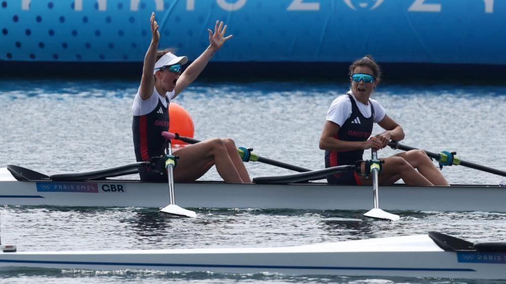 Emily Craig (left) with both her arms in the air with Imogen Grant, with her mouth open in surprise, after winning  a gold medal in a rowing boat. They both have sunglasses on.