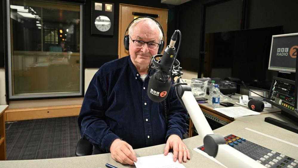 Paul Gambaccini wearing a dark blue shirt presenting a BBC Radio 2 programme. 