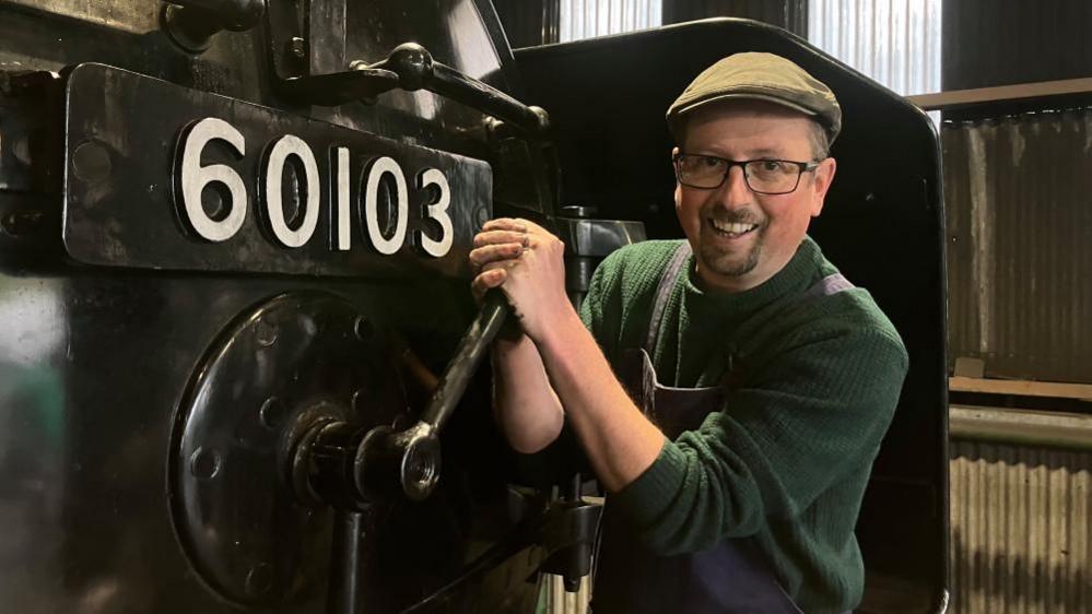 A white man with glasses and a short beard is standing right up alongside the front 'face' of a steam train. He holds a large metal crank in two hands and poses as if he is about to open the round metal door to the 'smokebox.' He wears a flat cap and blue dungarees over a green jumper. His hands are black with dirt or oil.