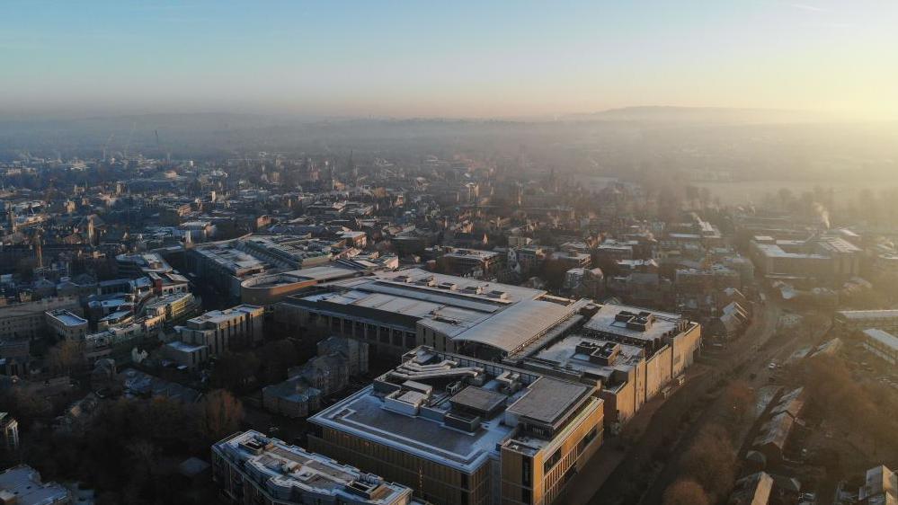 An aerial view of Oxford city on a winter morning. Spires are in the far distance, but it is mainly modern-looking buildings in the foreground.