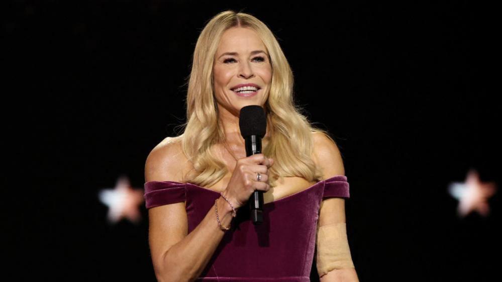 Chelsea Handler smiles as she addresses the audience on stage at the Critics Choice Awards. She is holding a black microphone up to her mouth and is wearing a purple dress