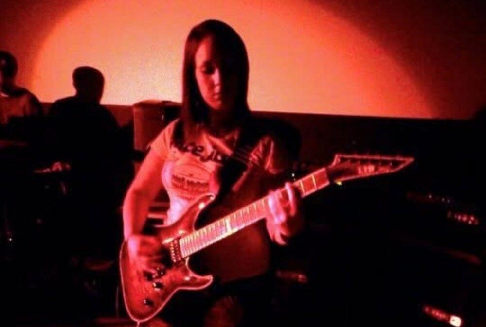 Sheree Gregg plays guitar on stage. The photo has a red filter. Sheree wears a t-shirt and has shoulder-length hair. She is looking down towards her guitar.