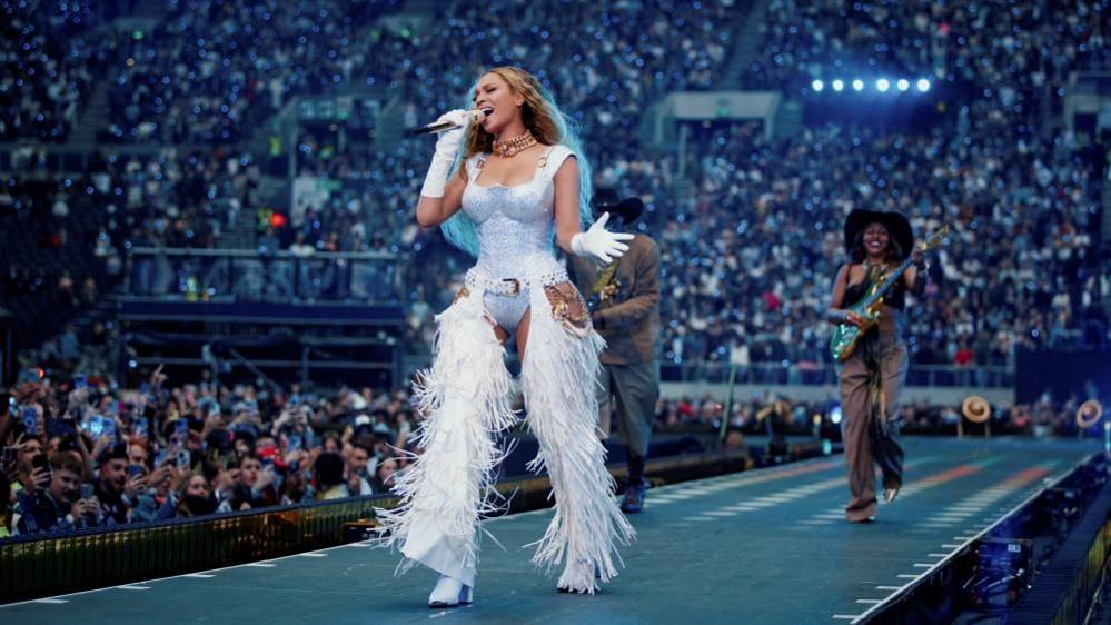 Beyoncé walks down a catwalk during a London concert wearing a white bodysuit and frilled chaps