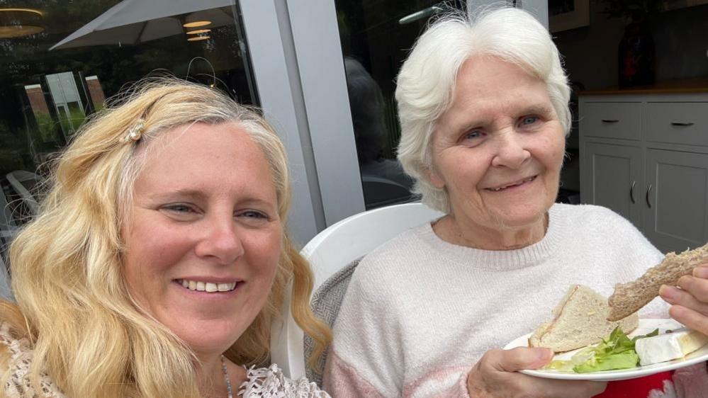 A selfie of a lady with blonde hair and an older lady to the right, who has grey hair and is holding a plate with sandwiches on. They are both smiling into the camera. 