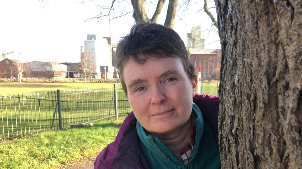 Marion Turner-Hawes with short black hair wearing a purple coat and green scarf, alongside a tree trunk.  There are black railings in the background, with a grass park behind and buildings beyond.