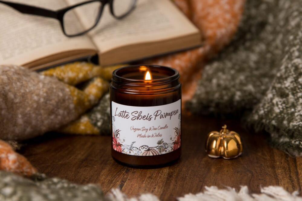 A cosy scene of a pumpkin spice candle, on a wooden table in front of a blanket, a book and a pair of glasses. It is next to a gold trinket in the shape of a pumpkin.