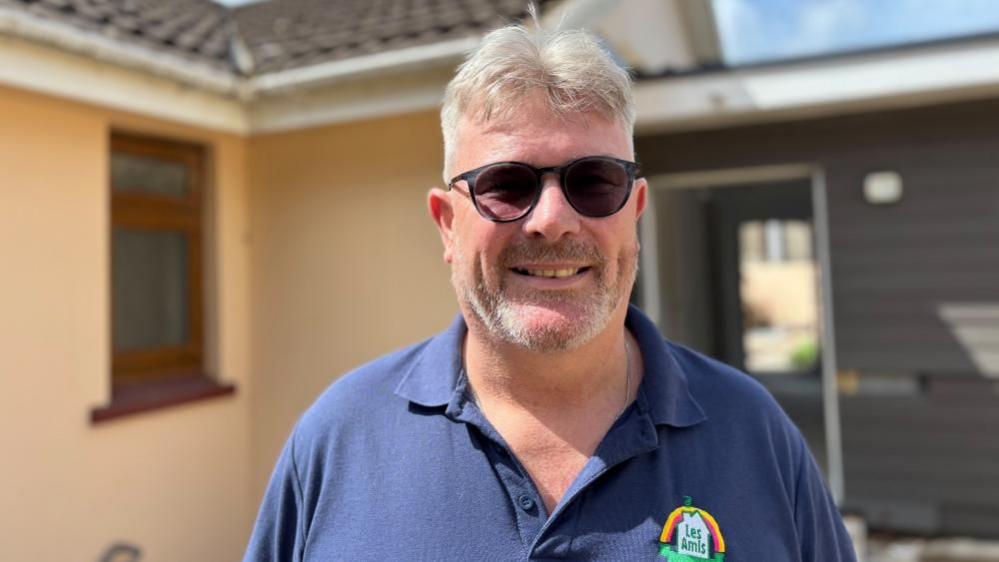 A man with grey hair and a beard, wearing sunglasses and a blue polo shirt with the Les Amis logo