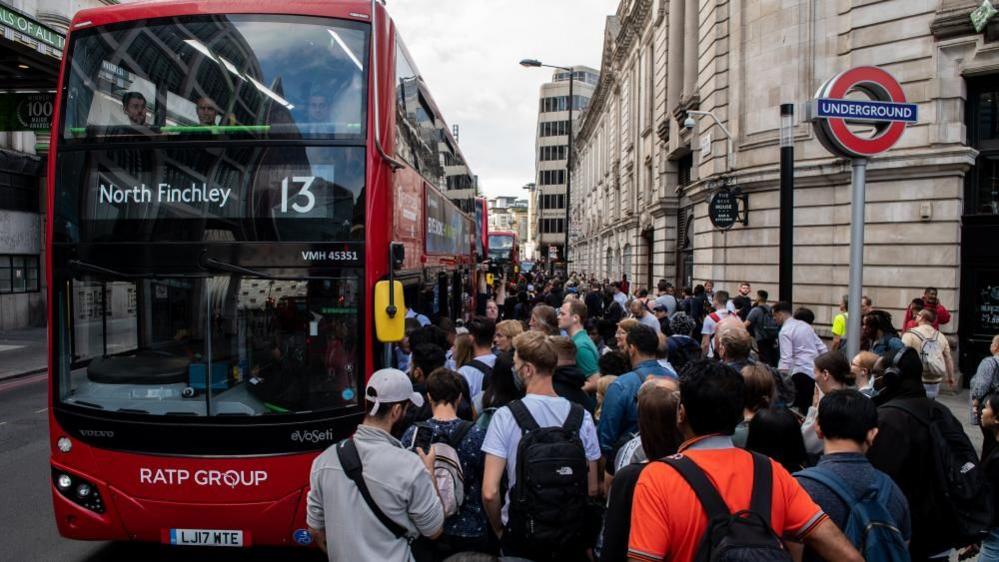 Commuters queue for buses outside Victoria underground train station. It is a busy day with a sea of people outside the bus. A red North Finchley 13 bus has arrived at the stop. 