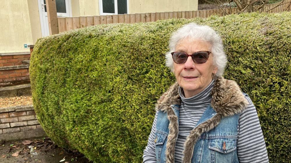 Gill Osborne, who is wearing a grey striped turtleneck jumper and a denim jacket with a fur collar, stands in front of a hedge. She has grey hair and is wearing sunglasses.