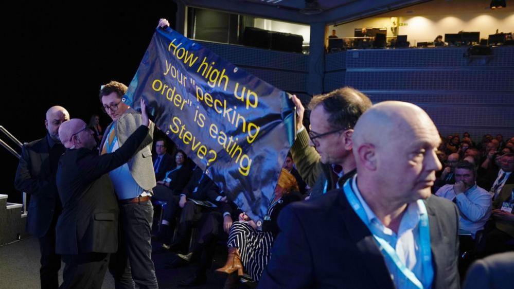 Protesters hold up a banner reading 'How high up your "pecking order" is eating Steve?' in the conference hall infront of Steve Reed