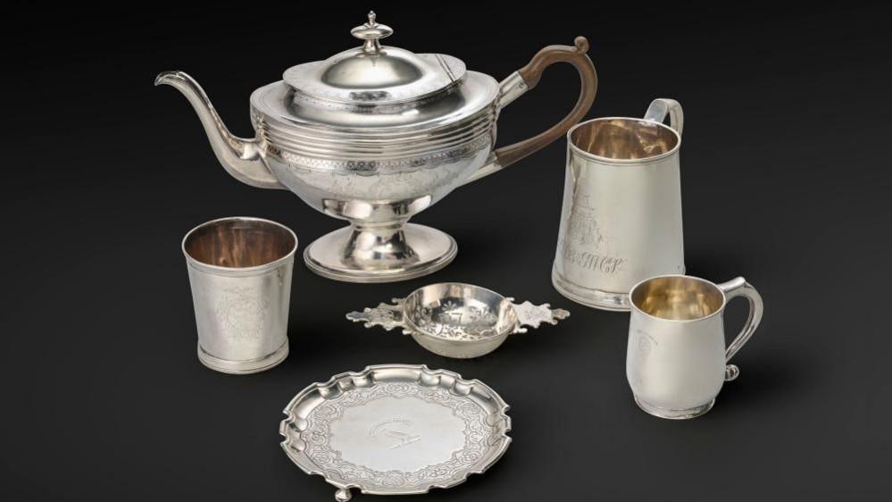 Silver items including a plate, jug, and three cups, against a dark background.
