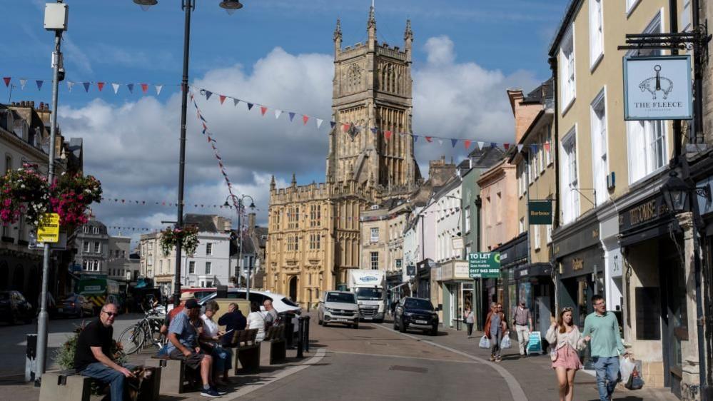 A road in central Cirencester with shoppers and bunting, leading up to the Parish Church of St John Baptist at Market Place
