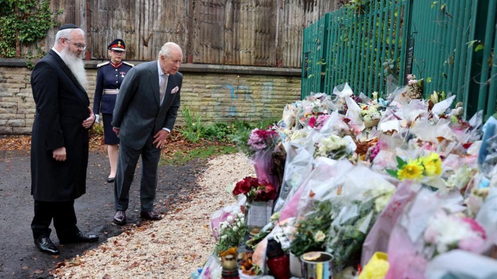 Prince Charles stands with a man with a long beard and wearing a kippah. They are both looking at bunches of flowers laid against a fence.
