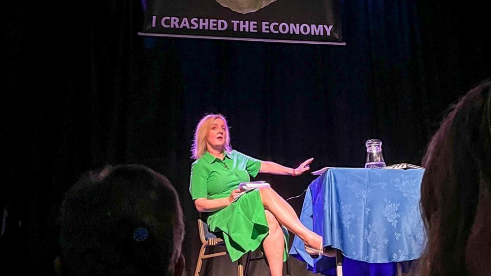 Liz Truss on stage with a blue cloth-covered table and a banner reading: "I crashed the economy"