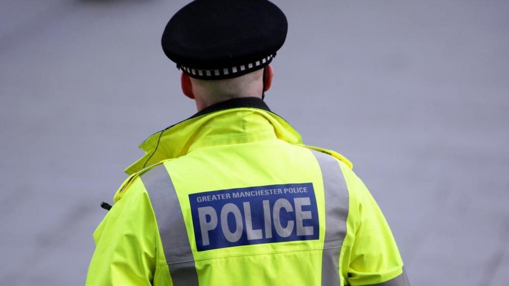 A police officer facing away from the camera wears a yellow high-vis jacket and a flat black police hat.
