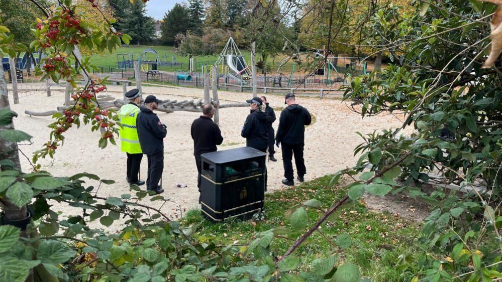 A group of police officers, five dressed in back, and one in a police hat, black trousers and a high-vis jacket, standing in a park, by a black bin, a playground, with trees and foliage around the site. There is sand on the ground. 