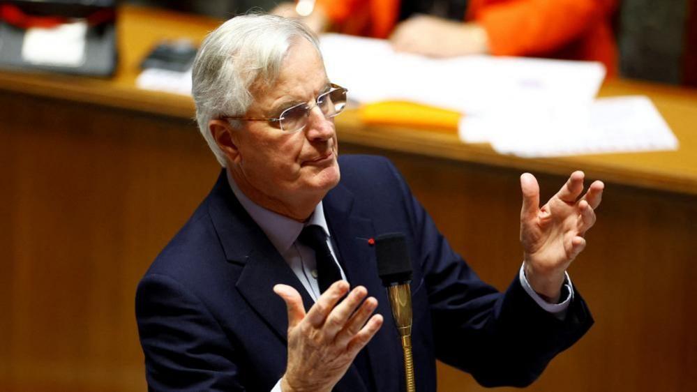 French Prime Minister Michel Barnier, wearing a dark suit jacket, shirt and tie, with his hands raised in front of him, speaks during the questions to the government session at the National Assembly in Paris