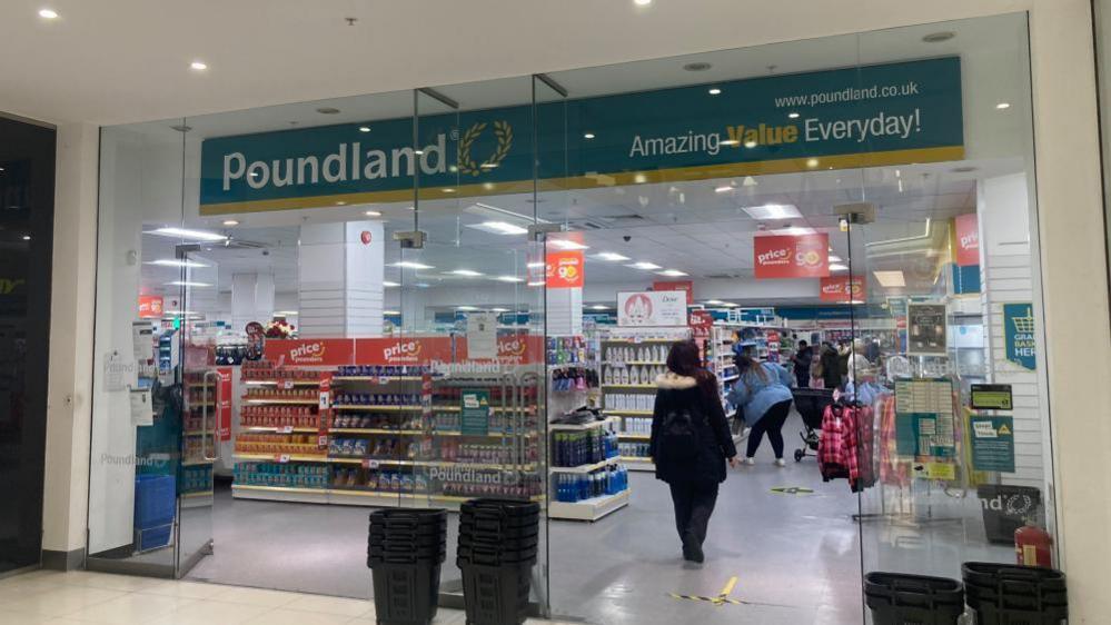 The shop front of a Poundland store inside a shopping centre. The store has glass windows and shoppers are just passed the entrance. There a black baskets by the doors. 