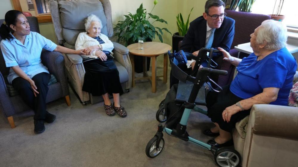 Andy Burnham is pictured sitting in a dark blue suit speaking to an elderly woman at a care home in north London. Another resident and a carer are pictured looking on.