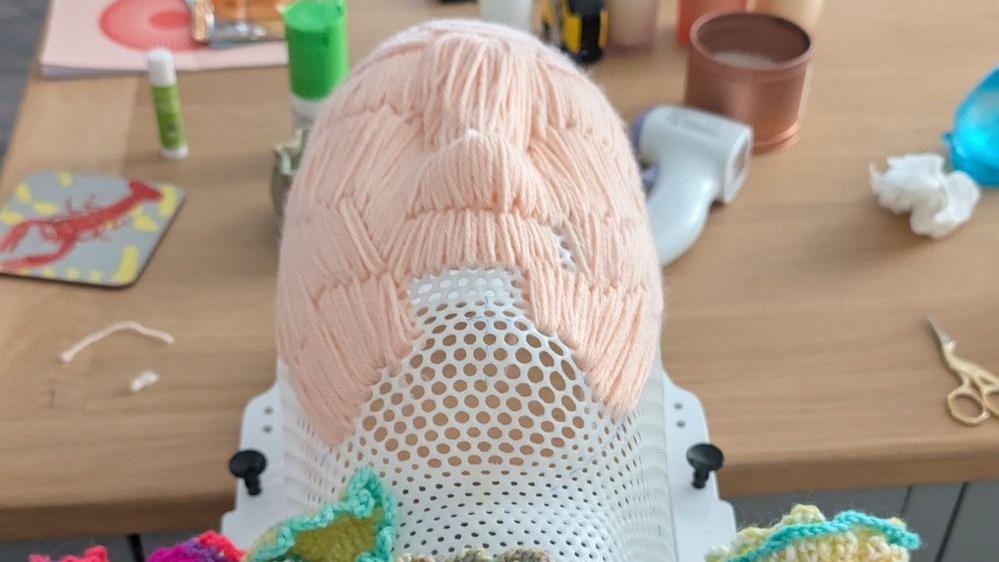 White plastic with many small holes in it, in the shape of a face. Coloured wool has been stitched through the holes in the mask.