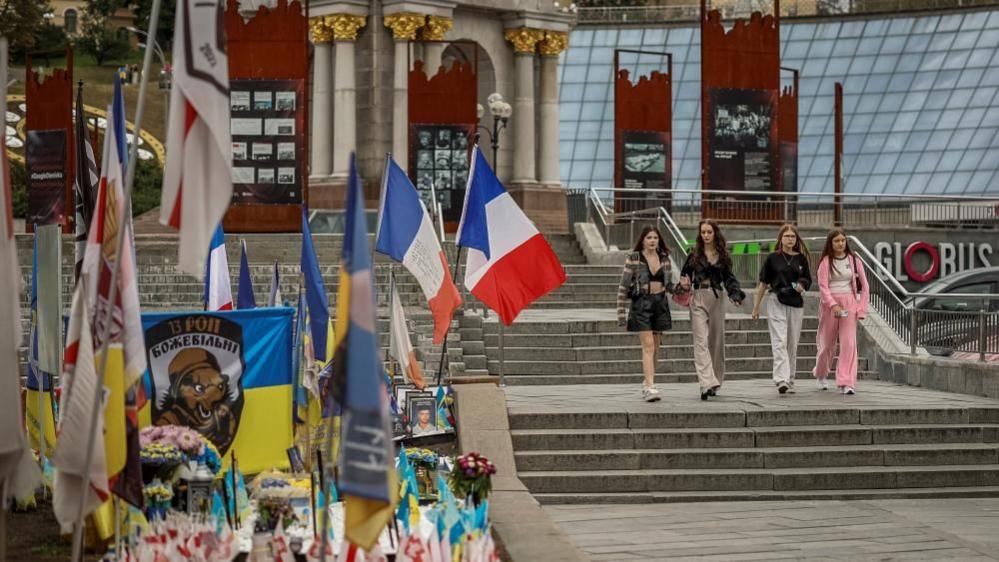 People pass by a makeshift memorial to fallen Ukrainian defenders in Kyiv's Independence Square. Along with flowers and the Ukrainian flag there are also flags of other countries, including France.