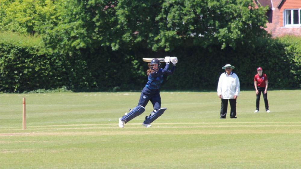 Libby uses her quick footwork to play the ball into the leg-side. Her right foot is on tip-toes whilst her left is planted, helping the ball around the corner towards  the square-leg umpire. Libby is wearing a navy blue cricket kit with matching helmet