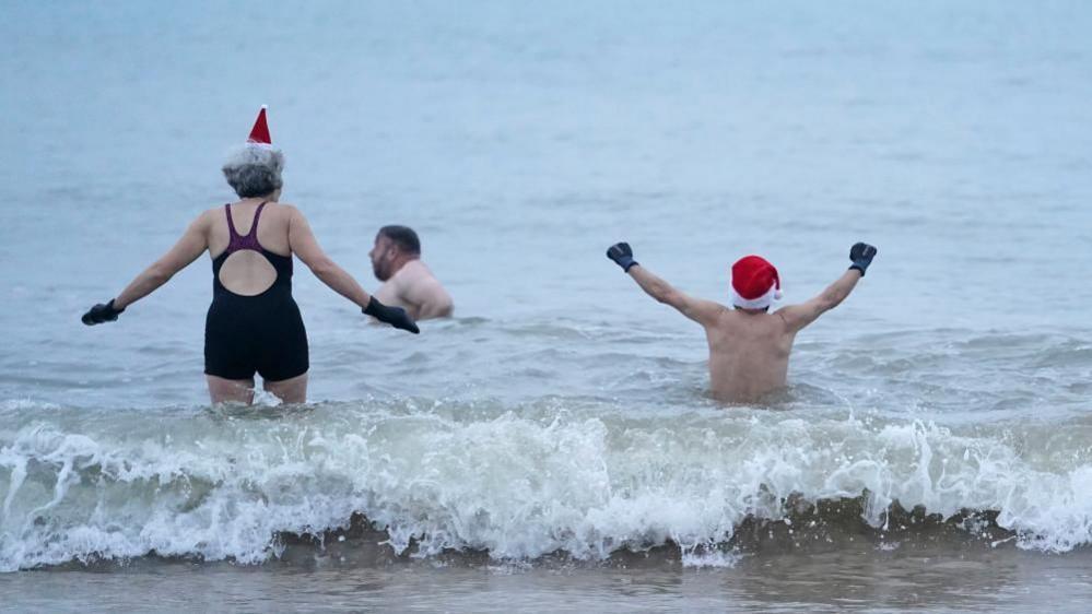 A woman in swimming costume with her back to us holds out her arms as though the sea she is wading into is bracing while a man, also with his back to us and wearing a santa hat, raises his arms in triumph