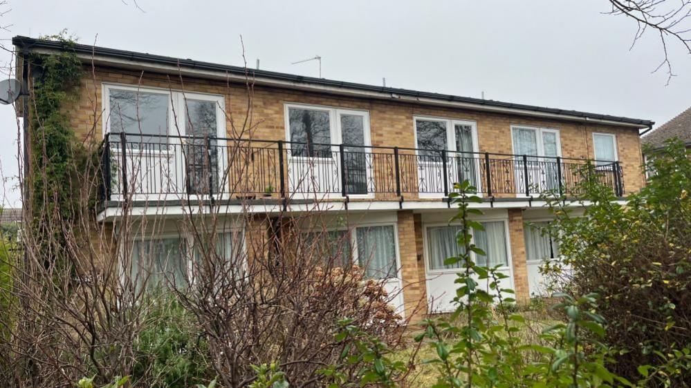 Abbeyfield House Care Home is a two-storey yellow brick building. It has large windows and a large balcony on its upper floor.  The building has a pitched tiled roof and hedging to the front.