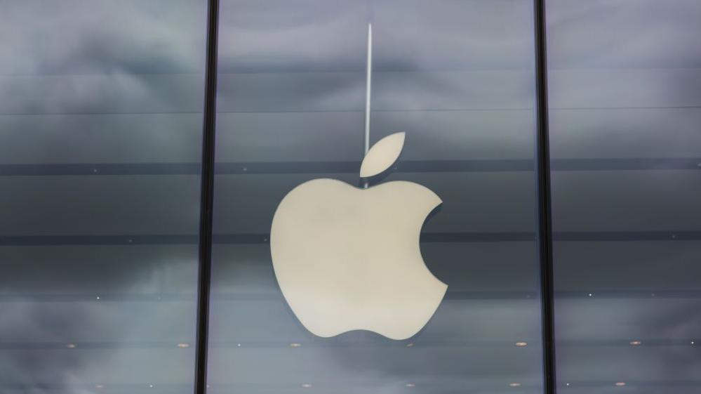 A white Apple logo on a glass fronted building, the glass reflects grey clouds.