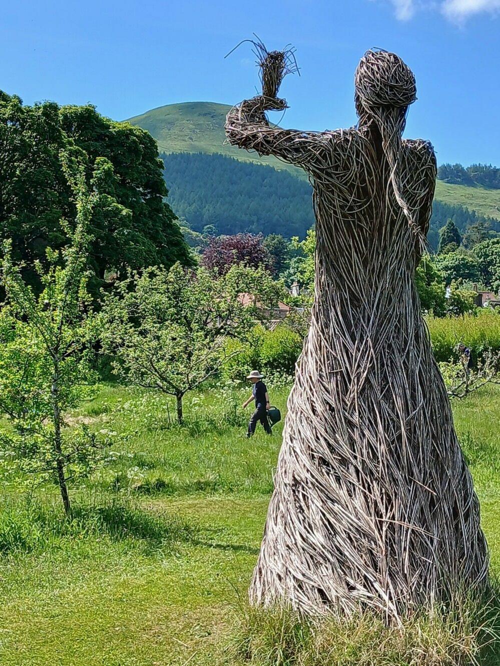 The back of a wicker figure looking towards bright green grassy hills and trees on a clear day.