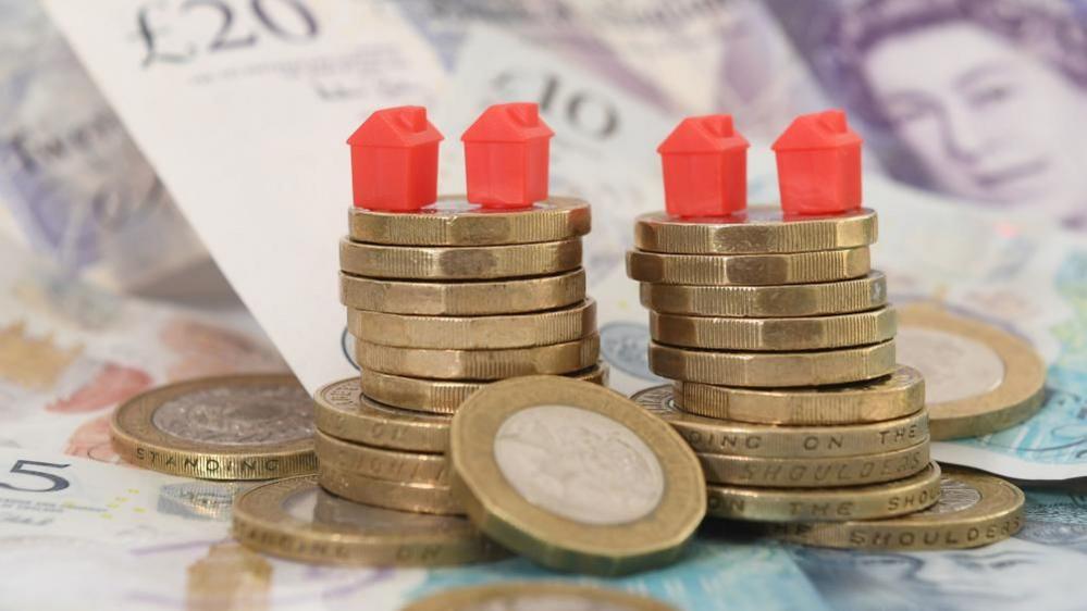 A stock image showing a pile of coins and notes, with four red mini model houses on top.