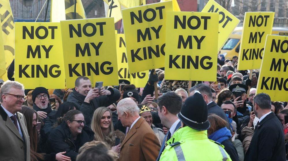 King Charles greeting the crowd of people at Centre Square. Large, yellow placards that read 'NOT MY KING' are being held up and cover the top of the photo.