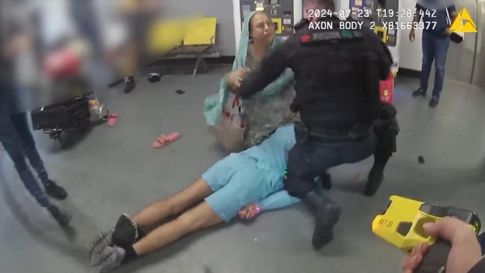 A screenshot from police bodyworn footage shows a man lying on his front on the floor. A woman and a police officer are both kneeling over him. A person's hand in the forearm of the shot is holding an electronic stun gun.