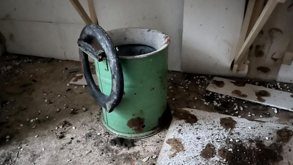 A green tin toilet with the toilet seat broken, hanging from the top of the hinge mechanism Debris and dirt is on the floor nearby.